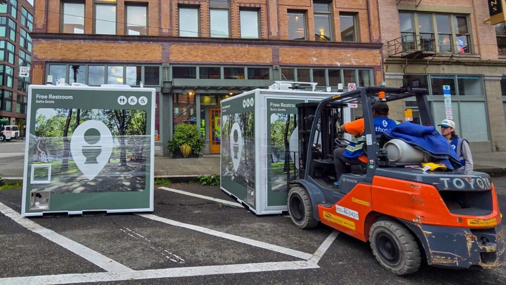 Trabajadores colocan módulos de baños públicos portátiles en un estacionamiento del centro de Seattle, frente a edificios comerciales de ladrillo. Un montacargas mueve una de las estructuras mientras continúa la instalación bajo un cielo nublado.