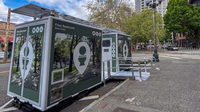 Módulos de baños públicos gratuitos instalados en una calle del centro de Seattle, cerca de la torre Smith Tower. Las estructuras incluyen acceso para personas con discapacidad y paneles solares en la parte superior.