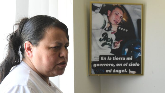 Adriana Garzón, Adriana Garzón, madre de Brayan Rayo Garzón, quien murió por suicidio mientras estaba bajo custodia del ICE en abril de 2025, posa junto a una foto de Rayo que dice 'En la tierra, mi guerrero; en el cielo, mi ángel', en el hogar de Garzón