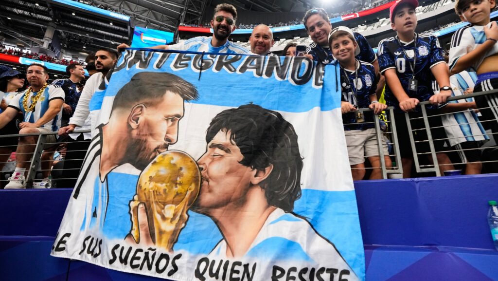Hinchas de Argentina sostienen una bandera con las imágenes de los astros argentinos Lionel Messi y Diego Maradona al término de la victoria 2-0 ante Austria en el Grupo J del Mundial, el lunes 22 de junio de 2026, en Arlington, Texas. (AP Foto/Tony Gutiérrez)