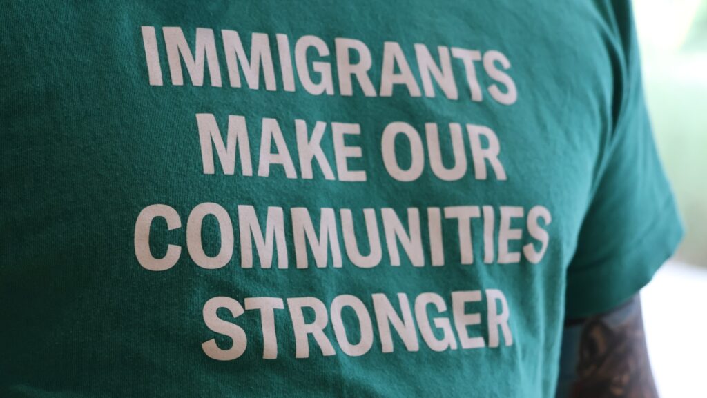 Primer plano de una camiseta verde con el mensaje en inglés “Immigrants make our communities stronger”, que significa “Los inmigrantes fortalecen nuestras comunidades”.