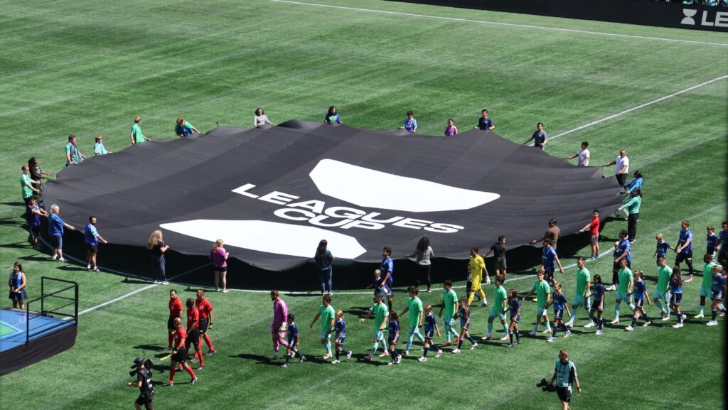 Jugadores de Seattle Sounders y Querétaro FC ingresan a la cancha de Lumen Field junto a una bandera de la Leagues Cup antes del partido.