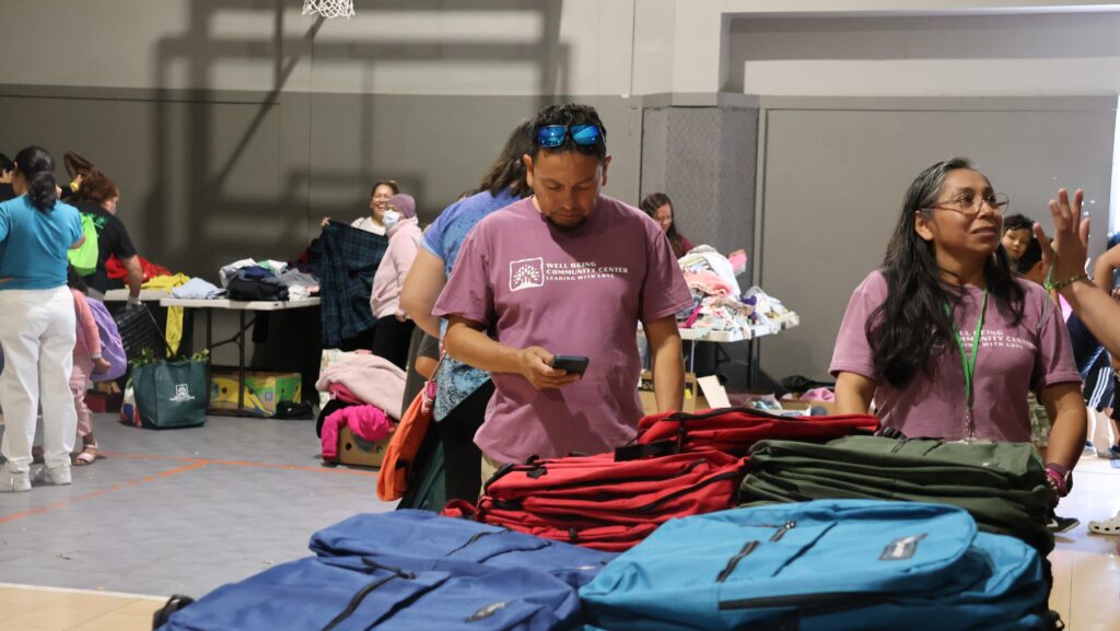 Voluntarios de Well Being Community Center junto a mochilas y ropa disponibles durante el evento de regreso a clases.