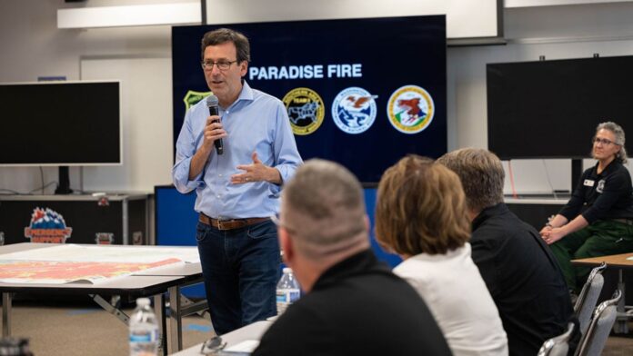 Bob Ferguson habla con un micrófono ante personal de emergencia, junto a un mapa y una pantalla con el nombre del incendio Paradise.