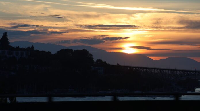 Julio rompe récord de calor en Estados Unidos mientras el humo amenaza regresar al Puget Sound Alt text: Atardecer sobre Seattle con el sol ocultándose detrás de las montañas Olímpicas, mientras una capa de neblina cubre parcialmente el horizonte del área de Puget Sound.