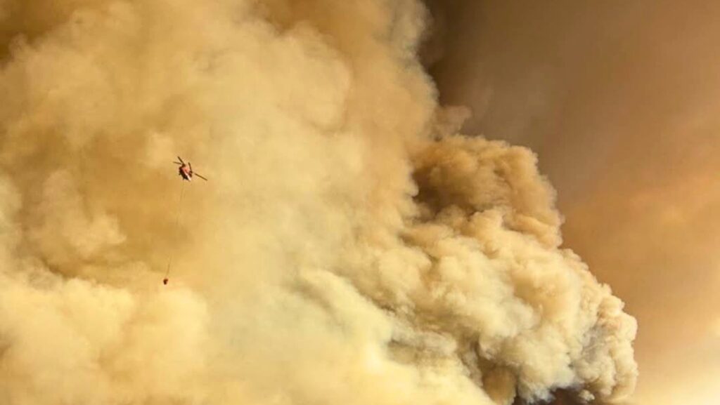Helicóptero realiza una descarga de agua mientras combate un incendio forestal cubierto por una densa nube de humo.