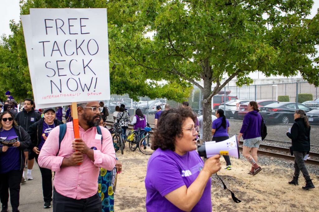 Manifestantes participan en una movilización frente al Centro de Detención de ICE en Tacoma mientras un asistente sostiene un cartel que exige la liberación de Tacko Seck.
