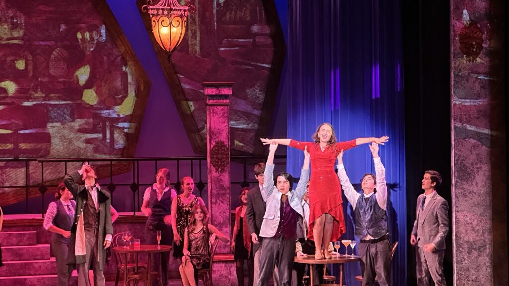 Maia Vazquez, con vestido rojo, interpreta a Lily durante una escena de Anastasia: The Musical en el Everett Performing Arts Center, rodeada por otros integrantes del elenco.