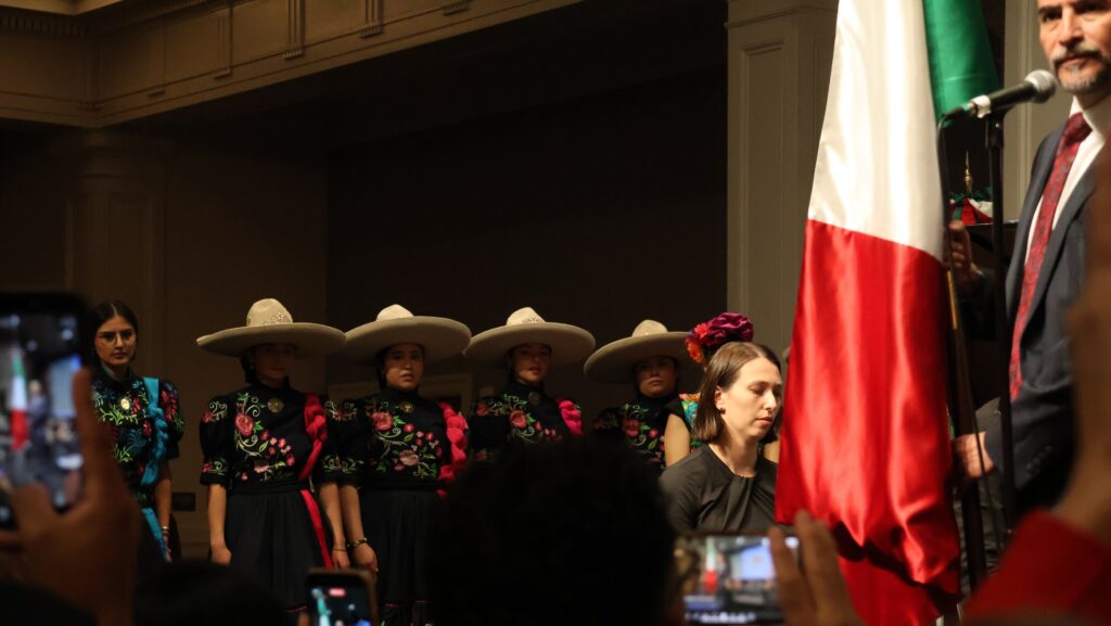 Víctor Hugo López Ortega frente a la bandera de México durante la ceremonia del Grito, acompañado por integrantes de la escolta.