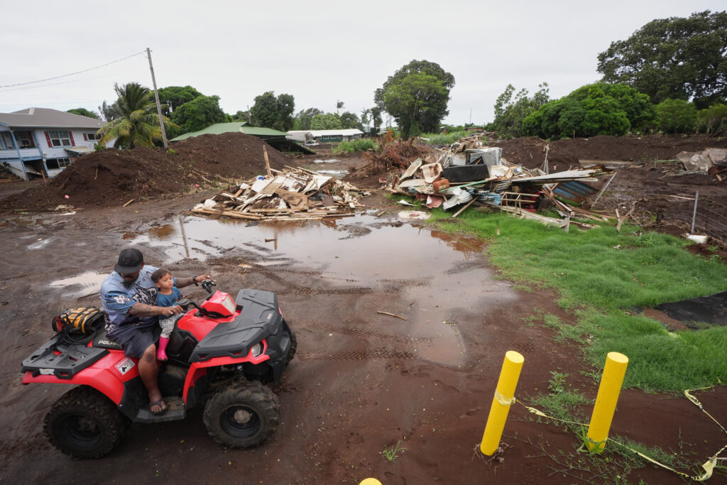 Hombre conduce un vehículo todoterreno con un niño frente a viviendas destruidas y escombros dejados por las inundaciones provocadas por el huracán Nolo en Naalehu, Hawái.