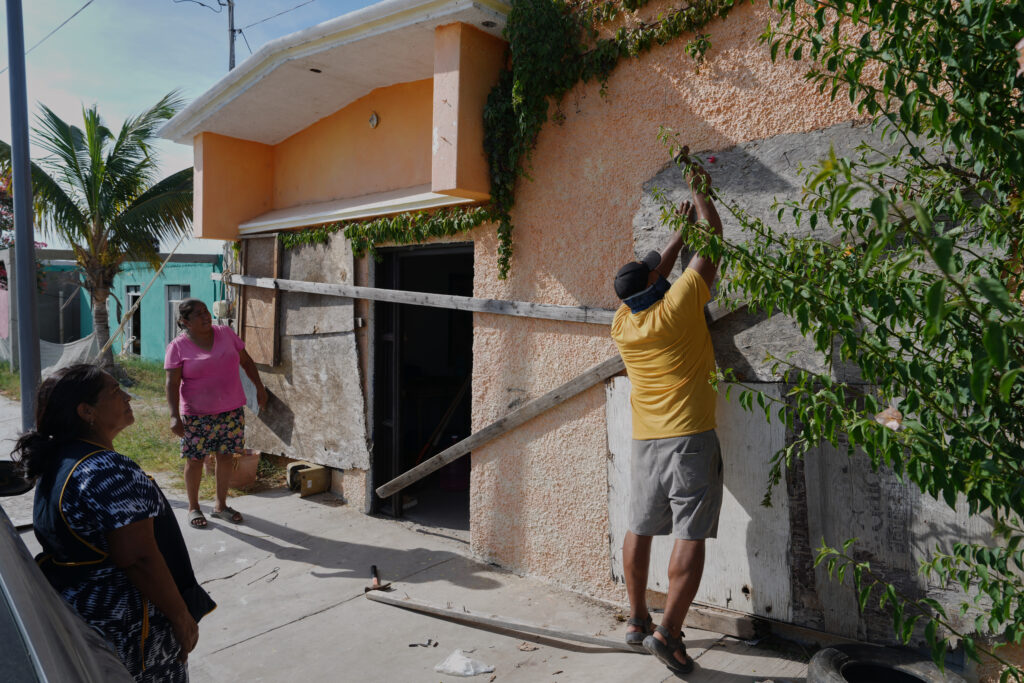 Hombre coloca tablones de madera sobre las ventanas de una vivienda mientras dos mujeres observan, ante la llegada del huracán Polo a Puerto San Carlos, Baja California Sur.