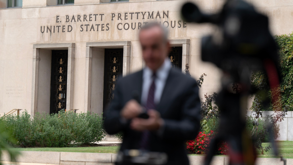 Fachada del E. Barrett Prettyman U.S. Federal Courthouse en Washington, D.C., mientras un hombre aparece frente a una cámara de televisión en primer plano.