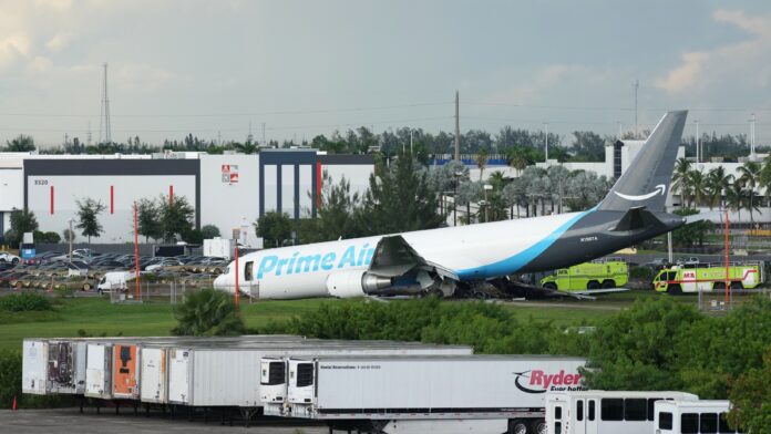 Avión de carga Prime Air de Amazon detenido fuera de la pista del Aeropuerto Internacional de Miami tras el accidente, mientras vehículos de emergencia permanecen junto a la aeronave.