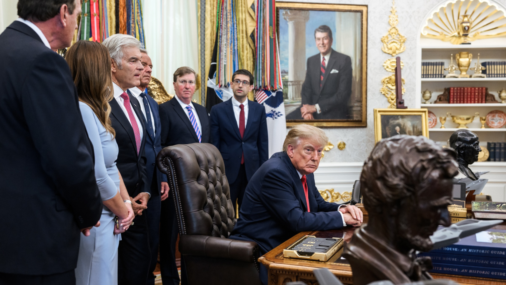 El presidente Donald Trump sentado en el escritorio de la Oficina Oval de la Casa Blanca, acompañado por varios funcionarios durante un evento.