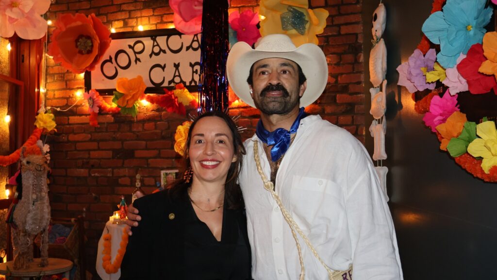 Sergio Garica y Karla Nahmmacher, organizadores del Día de Muertos en Pike Place Market, posan juntos en el restaurante Copacabana de Seattle, rodeados de flores de papel y decoraciones tradicionales. Garcia lleva un sombrero blanco y un pañuelo azul.