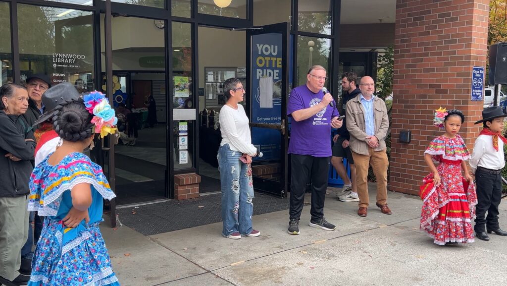 El alcalde de Lynnwood, George Hurst, habla ante los asistentes durante la celebración del Mes de la Herencia Hispana en Lynnwood Library, acompañado por Karina Gasperin y participantes con vestimenta tradicional.