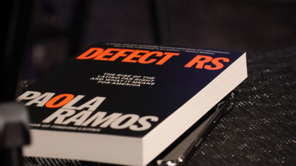 Primer plano del libro Defectors: The Rise of the Latino Far Right, de Paola Ramos, con una portada negra y letras blancas y anaranjadas, colocado sobre una mesa.