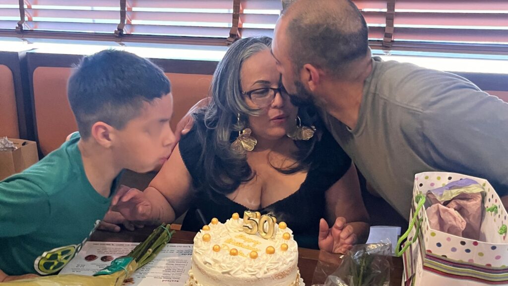 José Luis Arias besa en la frente a su esposa, Dania Salgado, durante la celebración de sus 50 años, mientras su nieto Mateo permanece a su lado frente a un pastel de cumpleaños.