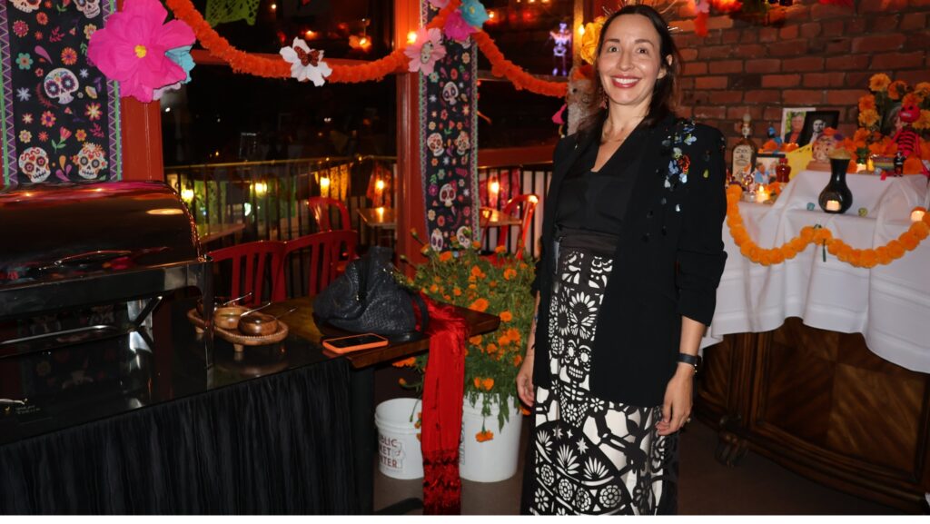 Karla Nahmmacher posa en el restaurante Copacabana, decorado con flores de papel, guirnaldas de cempasúchil y motivos del Día de Muertos. Detrás de ella se observa un altar con fotografías, velas y ofrendas.