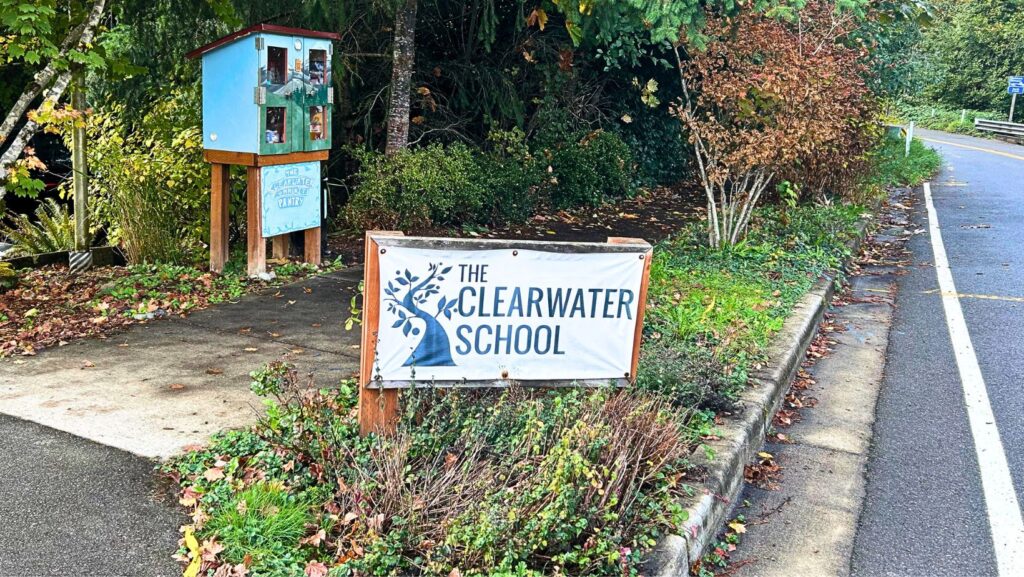 Letrero de The Clearwater School junto a una pequeña despensa comunitaria de madera, ubicada a un costado de la carretera en Bothell.