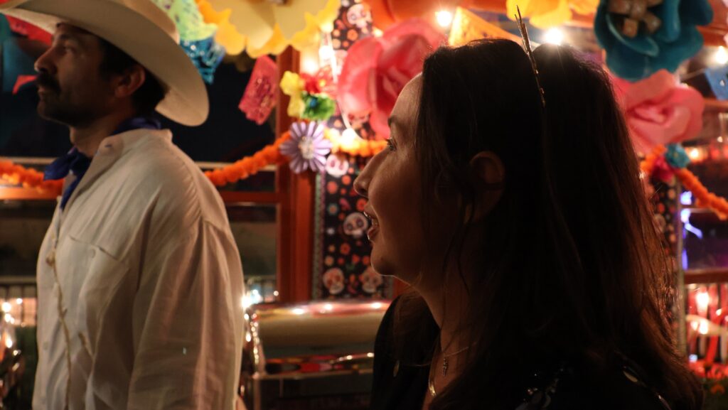 Sergio Garica, con sombrero blanco, camisa blanca y pañuelo azul, aparece junto a Karla Nahmmacher, vestida de negro, en el restaurante Copacabana, rodeados de flores de papel, luces y decoraciones tradicionales.
