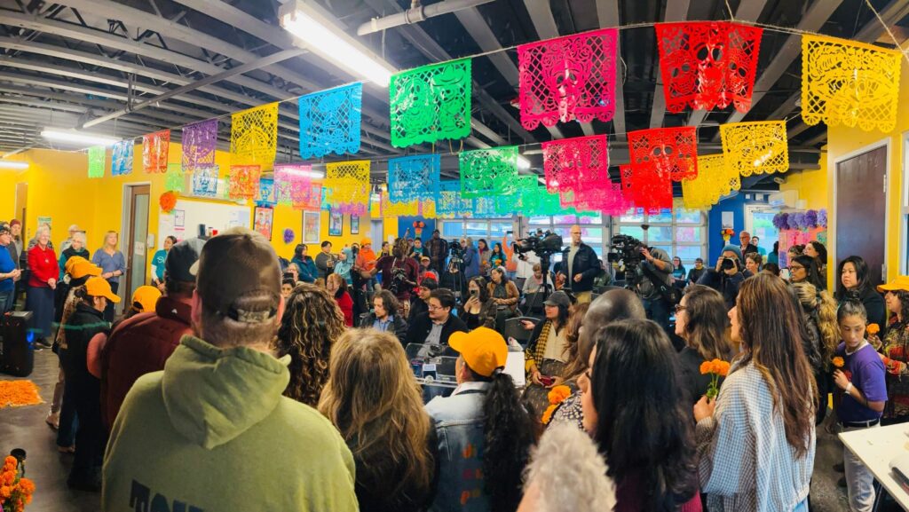 Decenas de personas y representantes de medios de comunicación participan en una reunión sobre operativos migratorios en Casa Latina, Seattle, bajo decoraciones multicolores de papel picado.