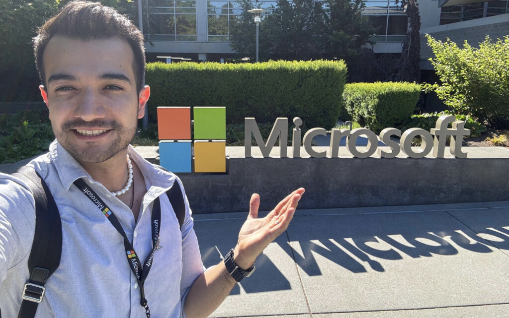 Carlos Iván Ibarra Pacheco sonríe y señala el logotipo de Microsoft frente a las instalaciones de la empresa durante su primer día como pasante, el 9 de junio de 2025.