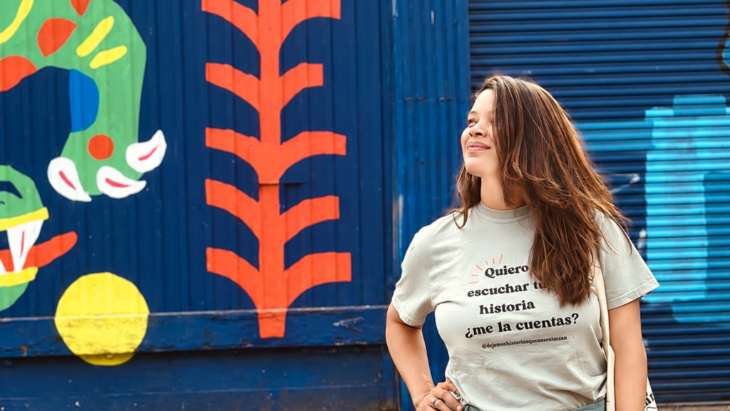 Sixta Morel posa frente a un mural de colores azul, verde, naranja y amarillo. Viste una camiseta blanca con el mensaje «Quiero escuchar tu historia, ¿me la cuentas?».