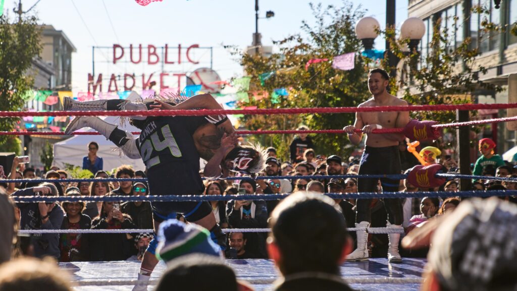 Un luchador enmascarado de Lucha Libre Volcánica levanta a su oponente para ejecutar un suplex en un cuadrilátero instalado en Pike Place Market. Otro luchador observa desde una esquina mientras numerosos espectadores rodean el ring.