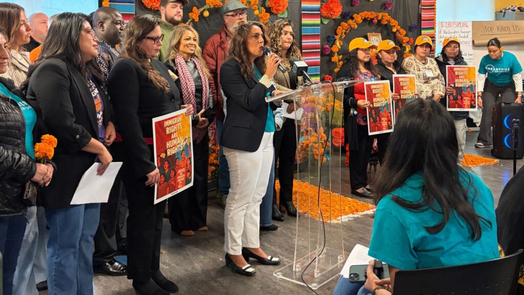 Una mujer habla ante un micrófono desde un podio transparente, acompañada por representantes de organizaciones defensoras de inmigrantes que sostienen carteles sobre derechos humanos en Casa Latina, Seattle.
