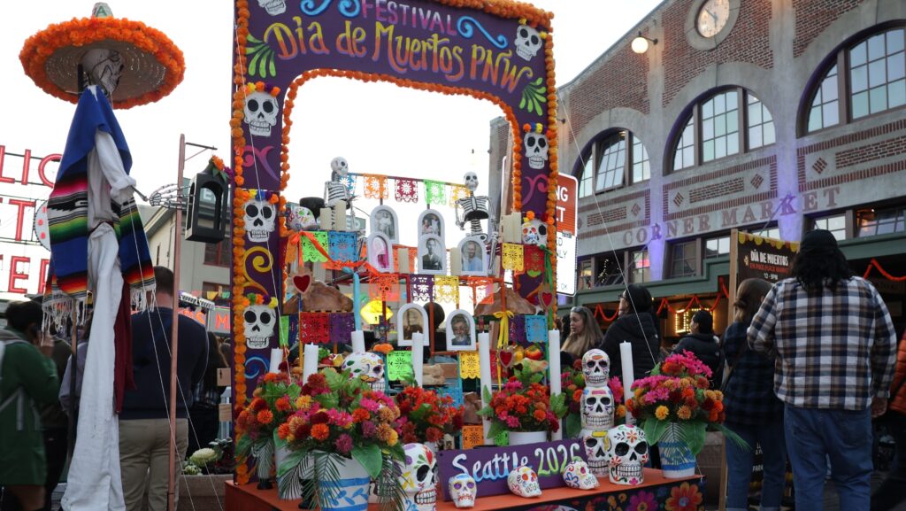 Altar principal del Día de Muertos instalado en la entrada de Pike Place Market, decorado con flores de cempasúchil, calaveras, velas, papel picado y fotografías de personas fallecidas. Un arco floral con el nombre del festival enmarca la ofrenda.
