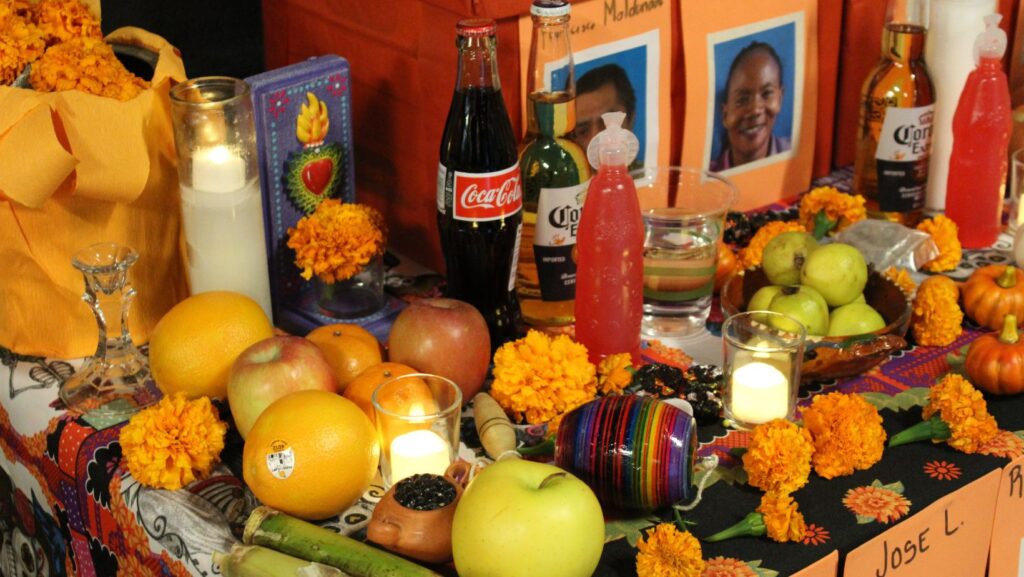 Primer plano de un altar de Día de Muertos con velas encendidas, flores de cempasúchil, frutas, bebidas, fotografías y objetos tradicionales durante una reunión comunitaria en Seattle.