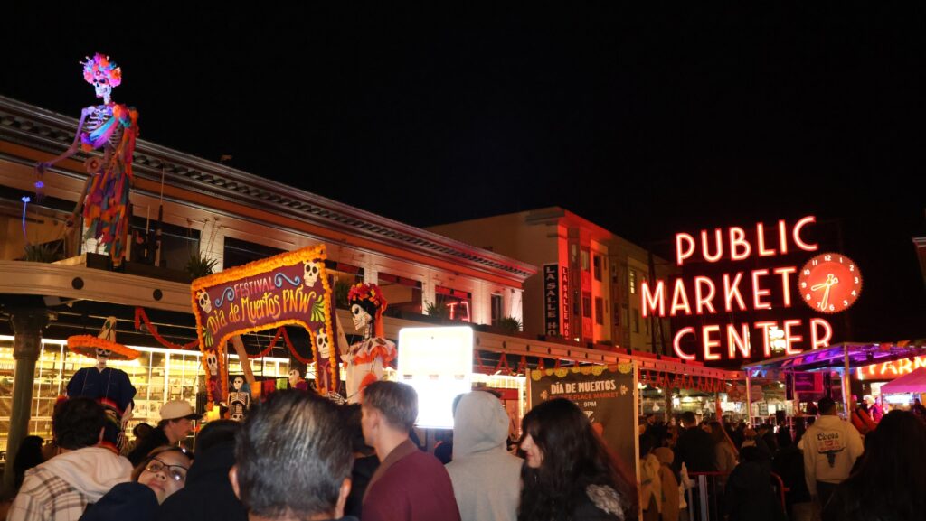 El altar del Día de Muertos y grandes figuras de esqueletos iluminados destacan frente al famoso letrero rojo de Pike Place Market durante la noche. Numerosos visitantes recorren el lugar entre decoraciones tradicionales y luces.