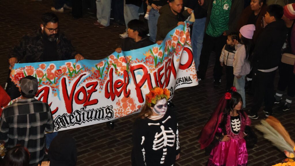 Participantes del desfile del Día de Muertos sostienen una pancarta colorida con las palabras «La Voz del Pueblo», mientras dos niñas caracterizadas con disfraces de esqueletos y maquillaje de calavera caminan por las calles de Pike Place Market.