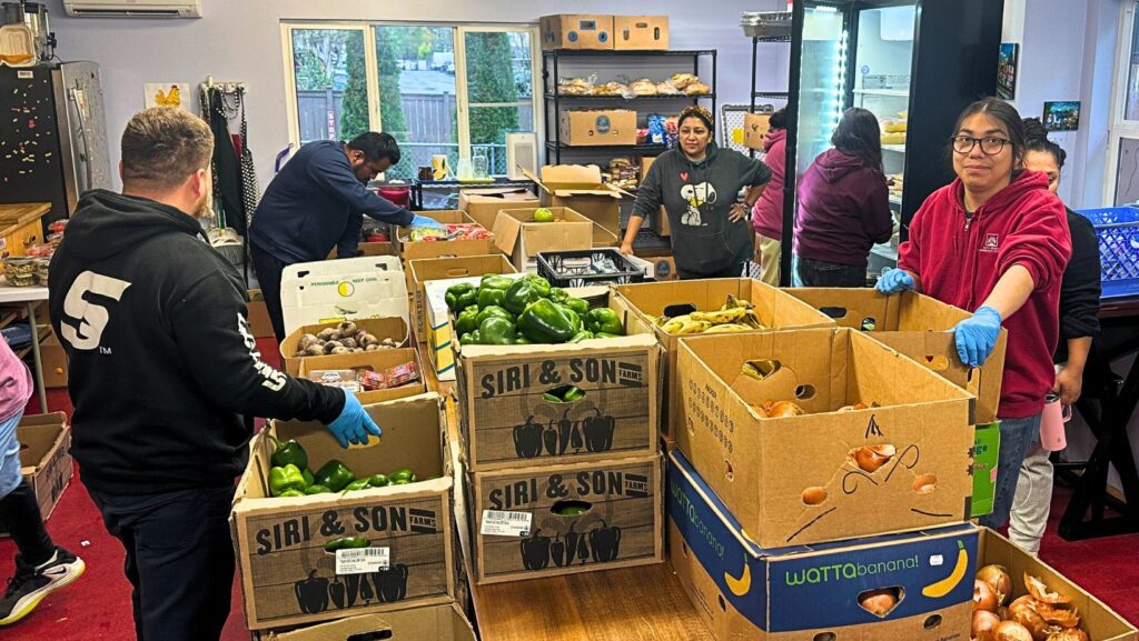 Voluntarios del Well Being Community Center trabajan entre cajas de pimientos verdes, cebollas, papas, plátanos y otros alimentos en The Clearwater School, Bothell.