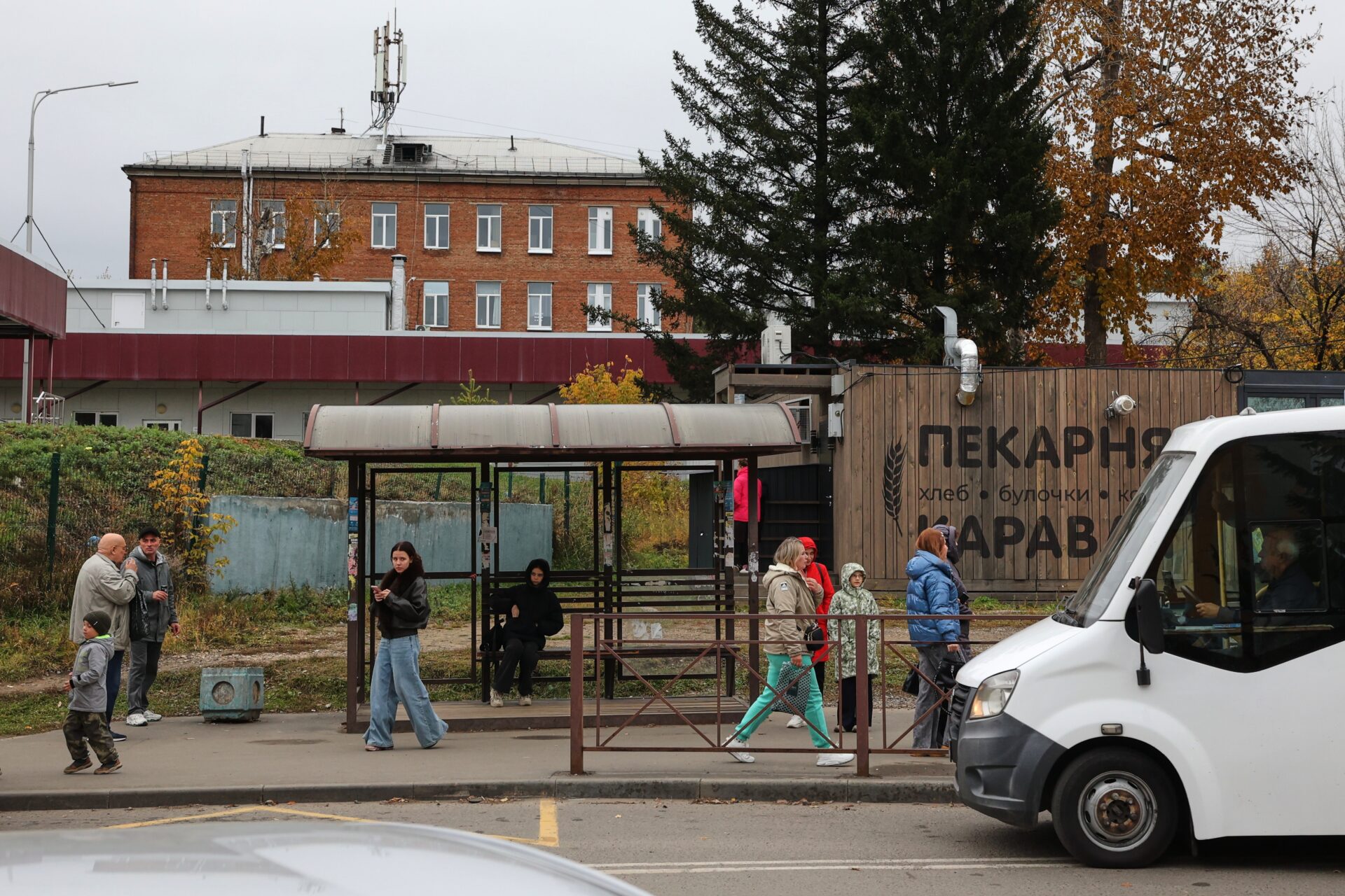 Personas esperan el transporte público y caminan frente a un edificio de ladrillo rojo en una calle de Rusia. En primer plano aparece una camioneta blanca y, al fondo, árboles con hojas otoñales.