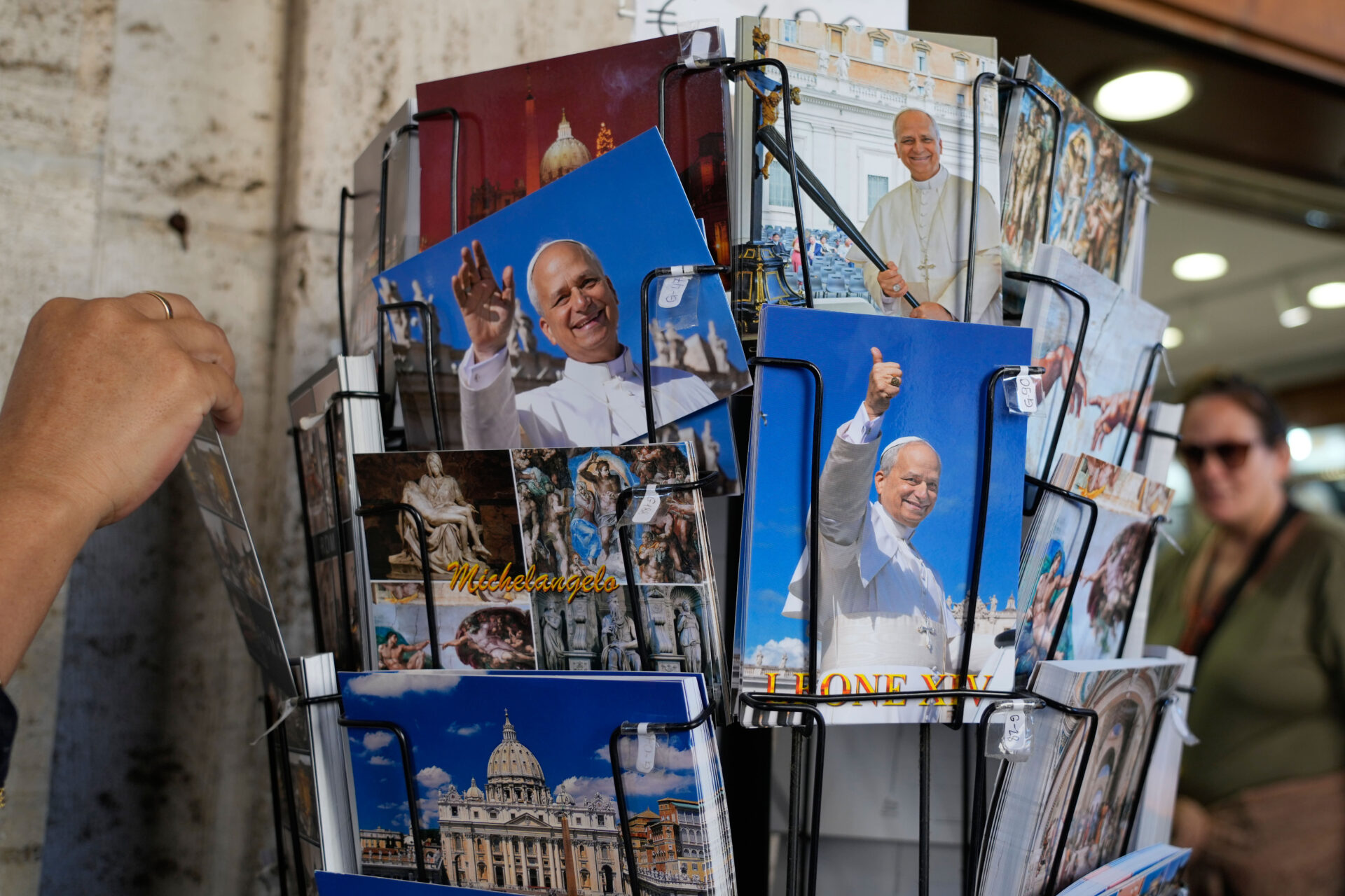 Exhibidor de postales y fotografías del papa León XIV en una tienda de recuerdos en Roma, Italia. Varias imágenes muestran al pontífice saludando y sonriendo, junto a postales de la Basílica de San Pedro y obras de arte religioso.