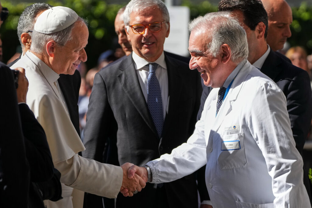 El papa León XIV estrecha la mano de un médico con bata blanca durante una visita al hospital Gemelli de Roma, Italia, mientras varios hombres observan el encuentro.