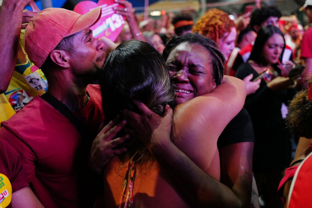 Simpatizantes de Luiz Inácio Lula da Silva se abrazan emocionados durante una celebración electoral en Brasil. Una mujer llora mientras otra la abraza, rodeadas de una multitud con ropa y artículos de campaña rojos.
