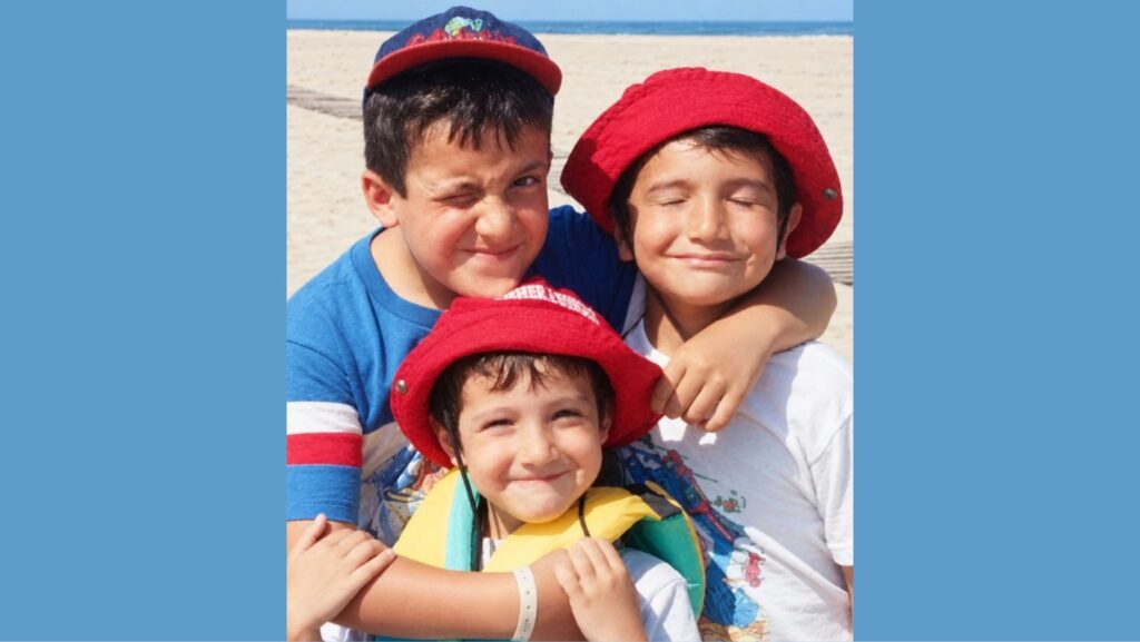 César, Cristian y Carlos Iván Ibarra posan abrazados durante su infancia en una playa. Los tres hermanos sonríen a la cámara y dos llevan sombreros rojos.