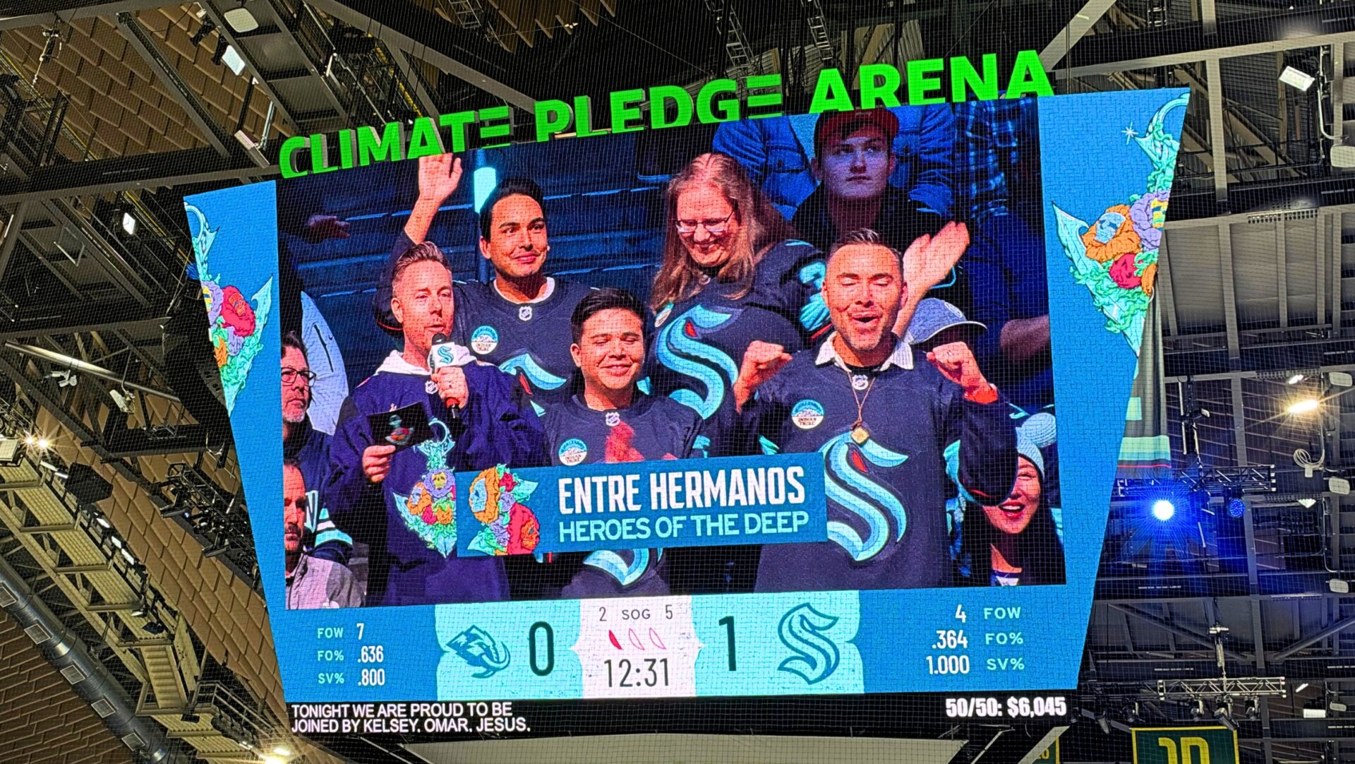 La pantalla gigante del Climate Pledge Arena muestra a integrantes de Entre Hermanos vestidos con camisetas de los Seattle Kraken durante la ceremonia de reconocimiento Hero of the Deep, mientras un presentador sostiene un micrófono.
