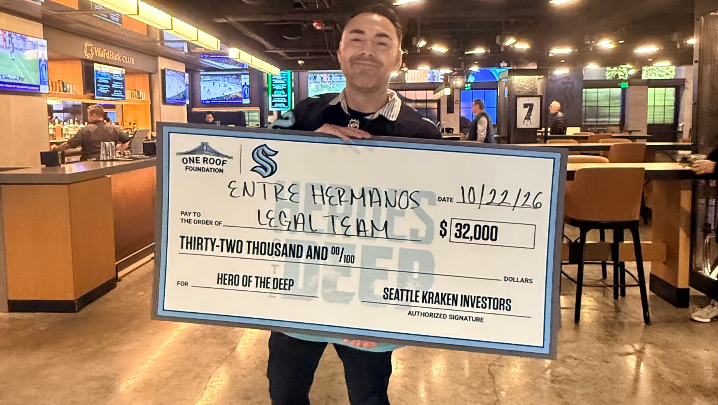Edgar Mack-Longoria sostiene un cheque simbólico de $32,000 otorgado al equipo legal de Entre Hermanos por los Seattle Kraken, durante una ceremonia en el Climate Pledge Arena.