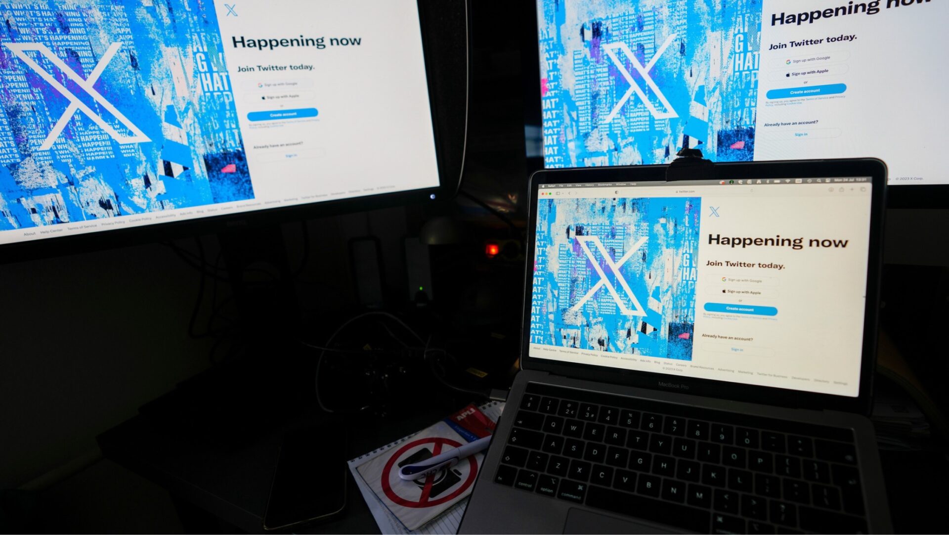 Tres pantallas de computadora, incluida una laptop, muestran la página de inicio de la red social X, anteriormente conocida como Twitter, con su logotipo blanco sobre un fondo azul y las opciones para crear una cuenta o iniciar sesión.