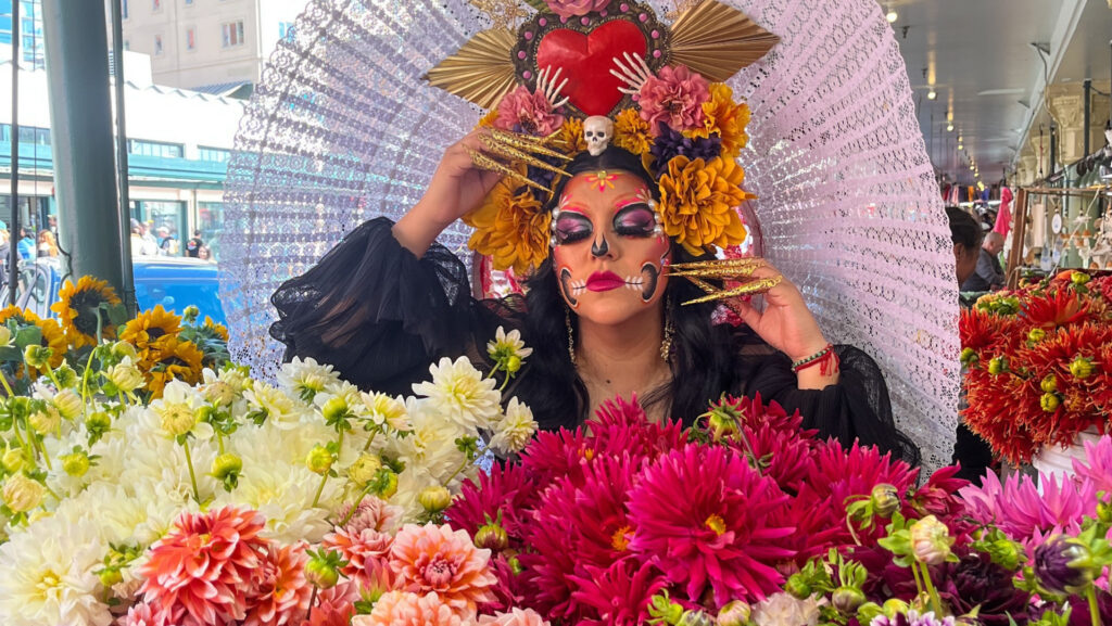 Mujer caracterizada como catrina, con maquillaje de calavera y un elaborado tocado de flores amarillas, posa entre arreglos florales en Pike Place Market, Seattle.