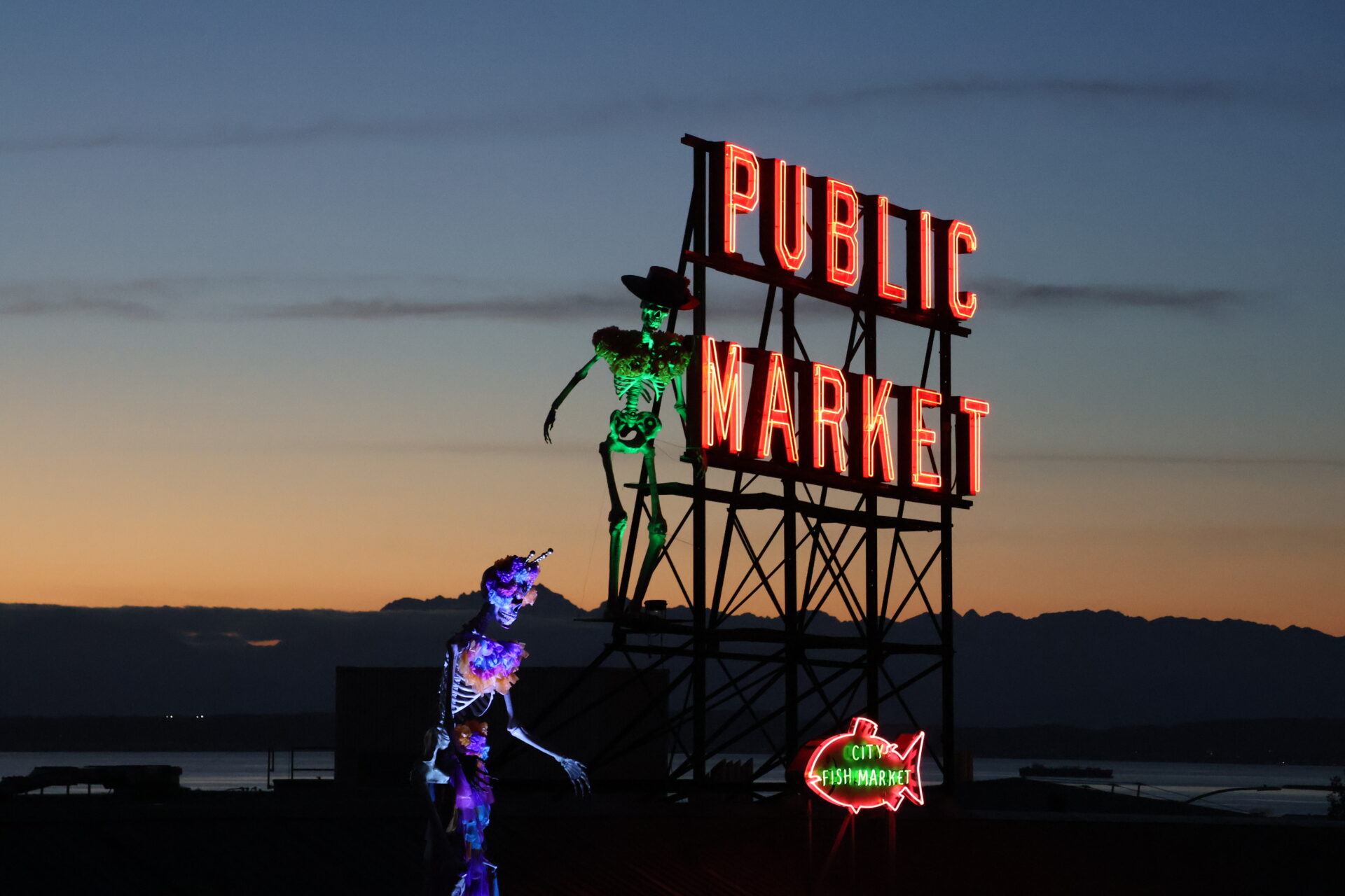 El letrero luminoso rojo de Pike Place Market destaca contra el cielo del atardecer en Seattle, acompañado de dos grandes figuras de esqueletos iluminados y decorados con sombreros y flores para la celebración del Día de Muertos.