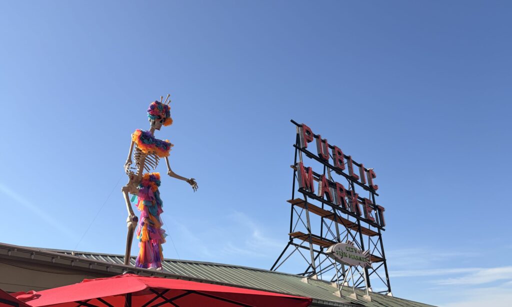 Figura gigante de una catrina esquelética, adornada con flores y telas de colores, junto al emblemático letrero de Pike Place Market en Seattle, bajo un cielo despejado.