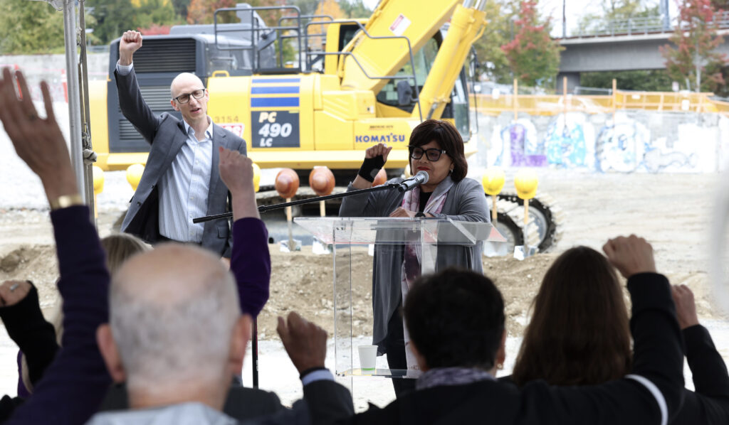 Estela Ortega, directora ejecutiva de El Centro de la Raza, habla frente a un micrófono durante la ceremonia de inicio de construcción del proyecto Mount Baker en Seattle, mientras los asistentes levantan los puños en señal de apoyo.