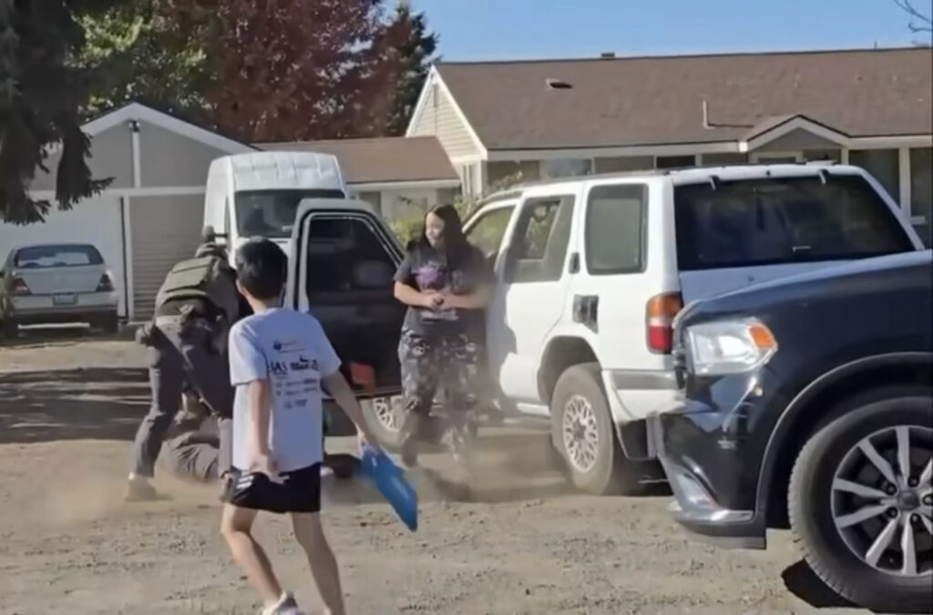 Un niño de siete años observa mientras dos personas vestidas de oscuro forcejean con un hombre en el suelo, junto a una camioneta blanca, durante un arresto en Spokane, Washington. Una mujer permanece cerca del vehículo.