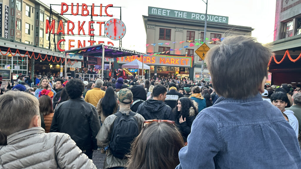 Una multitud llena la calle frente al escenario principal de Pike Place Market, en Seattle, durante la celebración del Día de Muertos. Al fondo se observa el famoso letrero luminoso del mercado y músicos sobre el escenario.