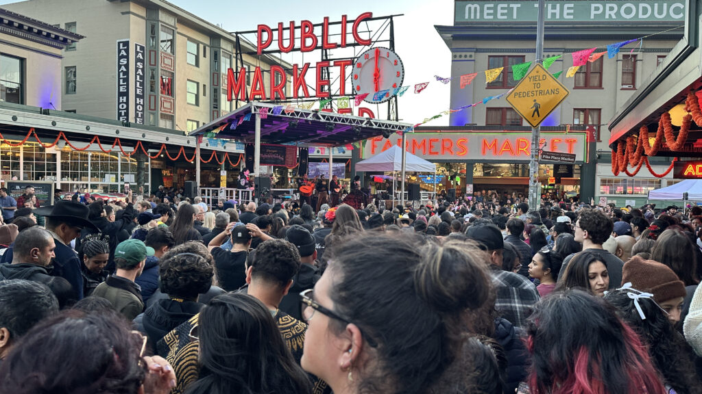 Miles de asistentes ocupan la calle frente al letrero de Pike Place Market durante las actividades del Día de Muertos en Seattle. Al fondo se observa un escenario con presentaciones artísticas y decoraciones de papel picado.
